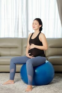 Happy fit pregnant young woman sitting on fitness ball concentrated on breathing and touching her belly with help of personal trainer